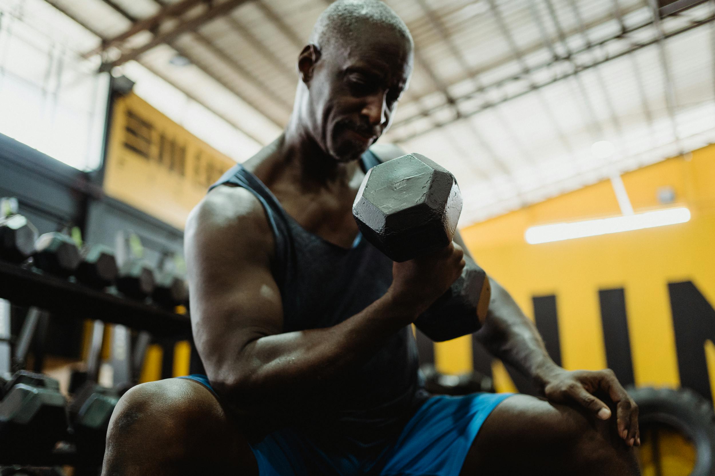 man wearing blue tank sitting in gym doing dumbbell concentration curl on bench